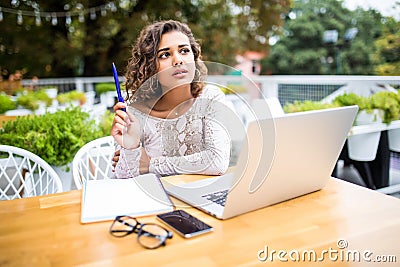 Photo of young excited latin curly woman indoors using laptop computer writing notes Looking aside have an idea in cafe Stock Photo