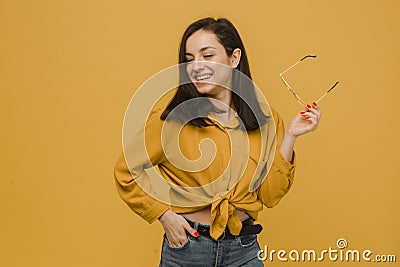 Photo of young female holds sunglasses and looks so happy. Wears yellow shirt, isolated yellow color background Stock Photo