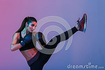 Photo of young sportswoman 20s in tracksuit and black boxing gloves fighting and kickboxing on training, isolated over dark Stock Photo