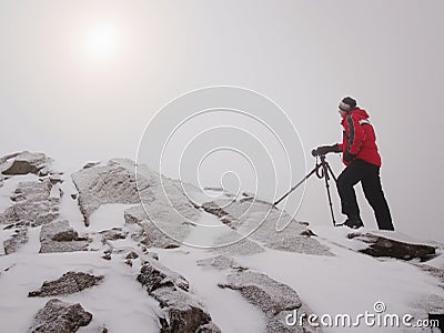 Photographer with eye at viewfinder of camera on tripod stay on snowy cliff and takes photos. Stock Photo