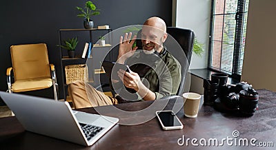 Photographer has online call with client. He holds Tablet PC and welcoming interlocutor. Desk in front equipped with Stock Photo