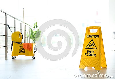 Phrase CAUTION WET FLOOR on safety sign and yellow mop bucket with cleaning supplies, indoors Stock Photo