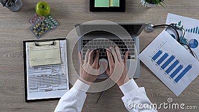 Physician looking at statistics chart, typing on laptop, preparing presentation Stock Photo