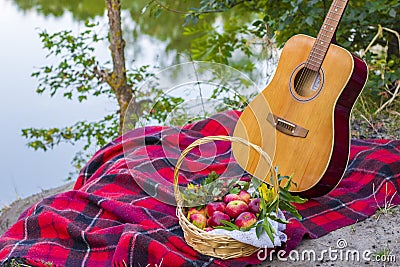 Picnic in nature. Picnic near river. Guitar and red apples in wicker basket on plaid blanket Stock Photo