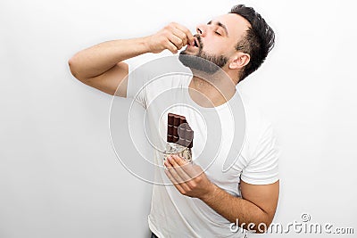 Picture full of pleasure. Young bearded man stands and eats chocolate. He puts piece of it into mouth. Guy keeps eyes Stock Photo