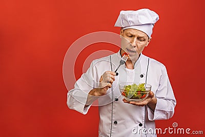 Picture of happy young senior chief cook in uniform standing isolated over red wall background, holding and eating salad Stock Photo