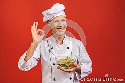 Picture of happy young senior chief cook in uniform standing isolated over red wall background, holding salad Stock Photo