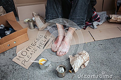 A picture of man`s feet without shoes. Man is sitting on cardboard. He is a beggar. There is a sign which says homeless Stock Photo