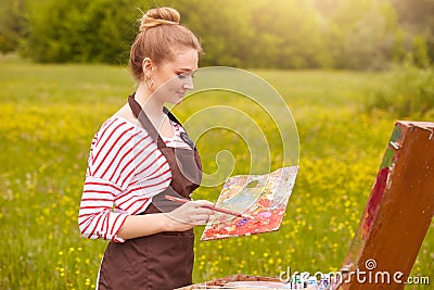 Picture of pleseant looking concentrated young Caucasian female with fair hair, artist standing in meadow with painting Stock Photo