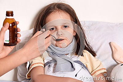 Picture of sleepy ill child drinking syrup, looking directly at camera, having tired look, lying in bed near her toy, unknown hand Stock Photo