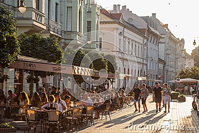 Young people walking on a pedestrian street of Szeged, Southern Hungary, with other people sitting on tables in cafes & restaurant Editorial Stock Photo