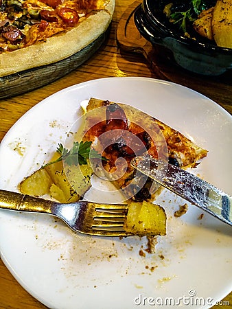 A piece of half-eaten pizza on a white plate with fork and knife. Wooden plate with a pizza and a bowl of baked potatoes Stock Photo