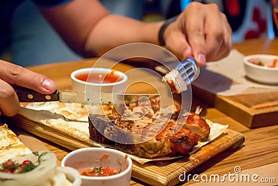 A piece of kebab, on a wooden plate, men`s hands holding a fork and a knife Stock Photo