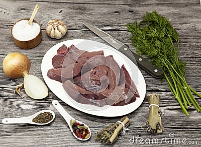Pieces of fresh raw beef liver, onion, garlic, spices, dill, parsley, salt, knife, olive oil on porcelain plate on a wooden backgr Stock Photo