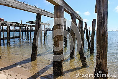 Piers from destroyed dock standing crooked out in the ocean with moss and barnacles around bottoms and a marina and sailboats in t Stock Photo