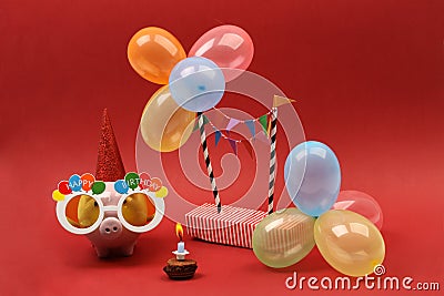 Piggy bank with sunglasses Happy birthday, party hat and multicolored party balloons on red background Stock Photo