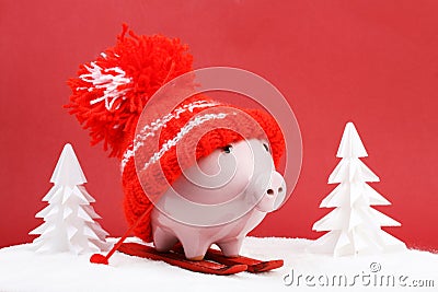 Piggy box with red hat with pompom standing on red ski and ski sticks on snow and around are snowbound trees on red background Stock Photo