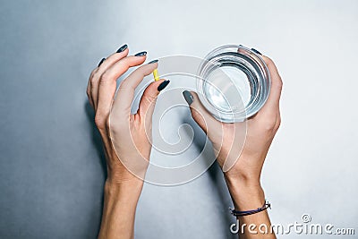 Hands of caucasian girl with yellow pill in left palm and glass of water in right hand. Stock Photo