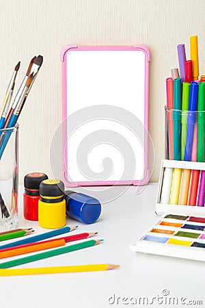 Pink digital tablet with a blank screen on a white table with multicolor drawing supplies. Stock Photo