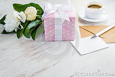 Pink dotted gift box, empty greeting card, kraft envelope, peonies bouquet and coffee cup over white wooden rustic table. Romantic Stock Photo