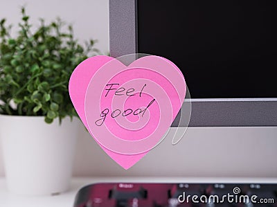 Pink heart shaped paper note with words Feel Good on it sticked on to a monitor at an office workplace Stock Photo