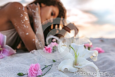 Pink lilies lying on sand foreground and young beatiful woman lying on sand at background on the beach at sunset Stock Photo