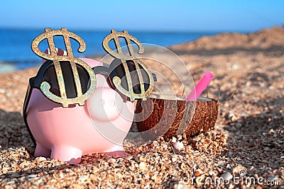 Pink piggy bank with sunglasses of dollar sings, standing on the beach sand close to coconut cocktail. Saving money for travel Stock Photo