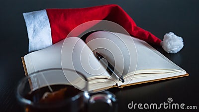 Planning future on Christmas concept: a notebook with blank pages, a black pen, Santa hat, glass mug with teabag, all on dark dini Stock Photo