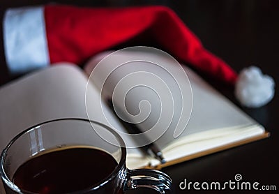 Planning future on Christmas concept: a notebook with blank pages, a black pen, Santa hat, glass mug with teabag, all on dark dini Stock Photo