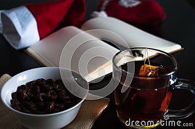 Planning future on Christmas: a notebook with blank pages, a black pen, Santa hat, glass mug with teabag, white bowl of chocolate Stock Photo