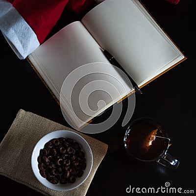 Planning future on Christmas: a notebook with blank pages, a black pen, Santa hat, glass mug with teabag, white bowl of chocolate Stock Photo