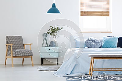 Plant on cabinet between patterned armchair and blue bed in bedroom interior with lamp. Real photo Stock Photo