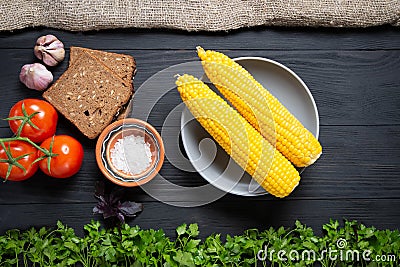Plate of corn on a black wooden background with lots of fresh vegetables around Stock Photo