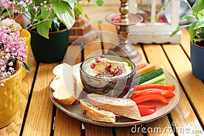 Plate with humus in a bowl decorated with pomegranate seeds, vegetable snacks, bread, on a wood table, selective focus. Stock Photo