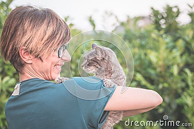 Playful domestic cat held and cuddled by smiling woman with eyeglasses. Outdoor setting in green home garden. Shallow depth of fie Stock Photo