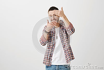 Playful embarrassed young boyfriend in trendy outfit and glasses covering face with palms, smiling broadly and being in Stock Photo