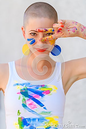 Playful portrait of a young gorgeous female artist painter covered in paint, looking and smiling at camera through her fingers. Stock Photo