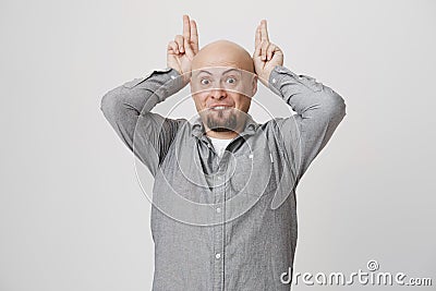 Playful positive bald male with beard has cheerful expression, wears gray shirt, behaves as rabbit, holds fingers above Stock Photo