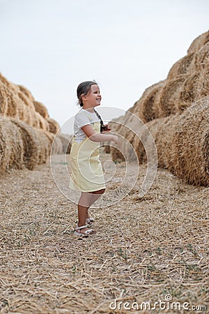 Playful shining little braided girl running on hay country field road between haystacks looking away in sundress. Having Stock Photo