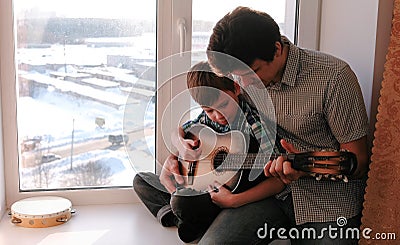 Playing a musical instrument. Dad teaches his son to play the guitar, sitting on the windowsill. Stock Photo