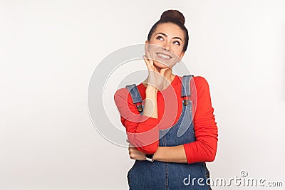 Pleasant memories. Portrait of positive inspired stylish girl with hair bun in denim overalls looking up with dreamy face Stock Photo