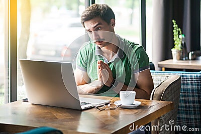 Please help me! Young worry businessman in green t-shirt sitting, working and looking at laptop display and begging to help or Stock Photo