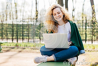 Pleased European woman having beautiful fluffy blonde hair wearing jeans, jacket and sport shoes sitting crossed legs using Stock Photo