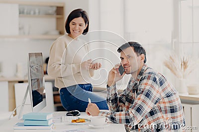 Pleased husband and wife manage finances at home, review bank accounts, pleased man holds mobile phone near ear, wears plait shirt Stock Photo