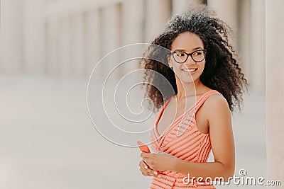 Pleased millennial girl with curly hair, wears pink dress, optical glasses, focused aside with smile, stands outdoor, waits for Stock Photo