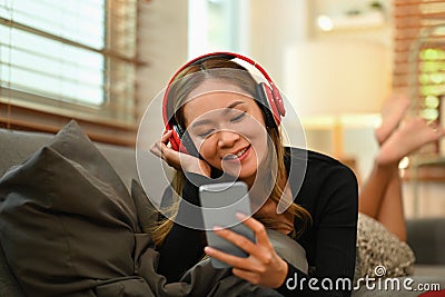 Pleased millennial woman with wireless headphone lying on comfortable couch and using smart phone Stock Photo