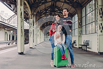 Pleased wife and husband cuddle with love, their daughter sits at suitcase, pose together on platform, look in ditance, wait for Stock Photo