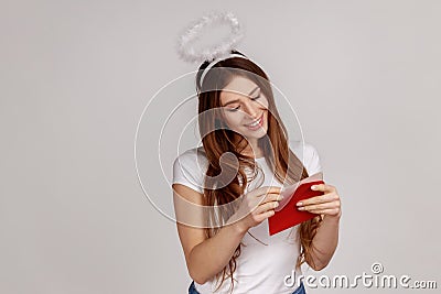 Pleased woman with nimbus over head holding red envelope, reading romantic letter with smile. Stock Photo