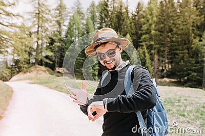 Pleased young man in sunglasses looking at wristwatch with smile standing on the road on nature background. Portrait of Stock Photo