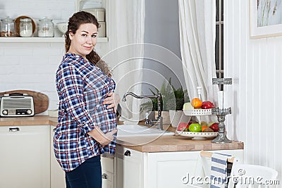 Pleasured pregnant woman holding her belly, dressed plaid shirt, curly long hairs. Female is in kitchen interior Stock Photo